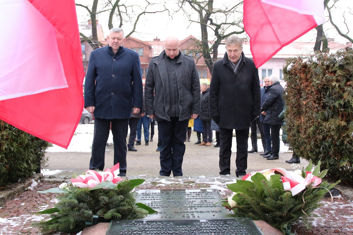 Delegacja miasta Sokołów Podlaski tuż po złożeniu kwiatów obok tablicy upamiętniającej. Władze miasta stoją w pozycji na baczność z pochylonymi głowami oddając hołd powstańcom. Na bokach widać biało - czerwone flagi.