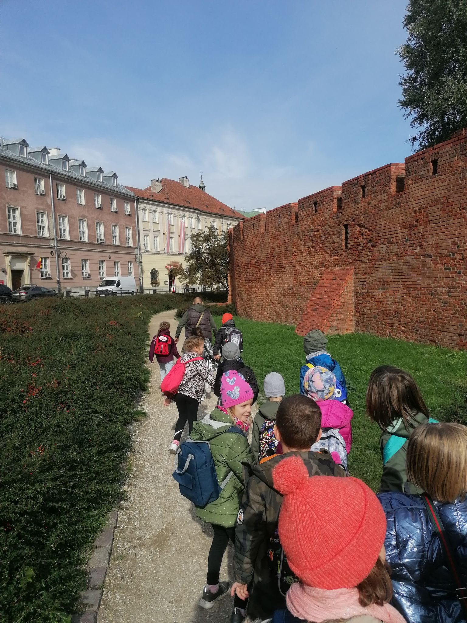 Dzieci idące chodnikiem. Po prawej stronie widać mur z czerwonych cegieł