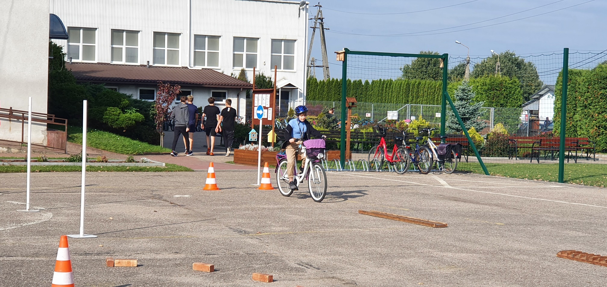 Dziewczynka w niebieskiej kurtce i niebieskim kasku jedzie rowerem po placu, gdzie trasę jazdy wyznaczają cegły i pachołki