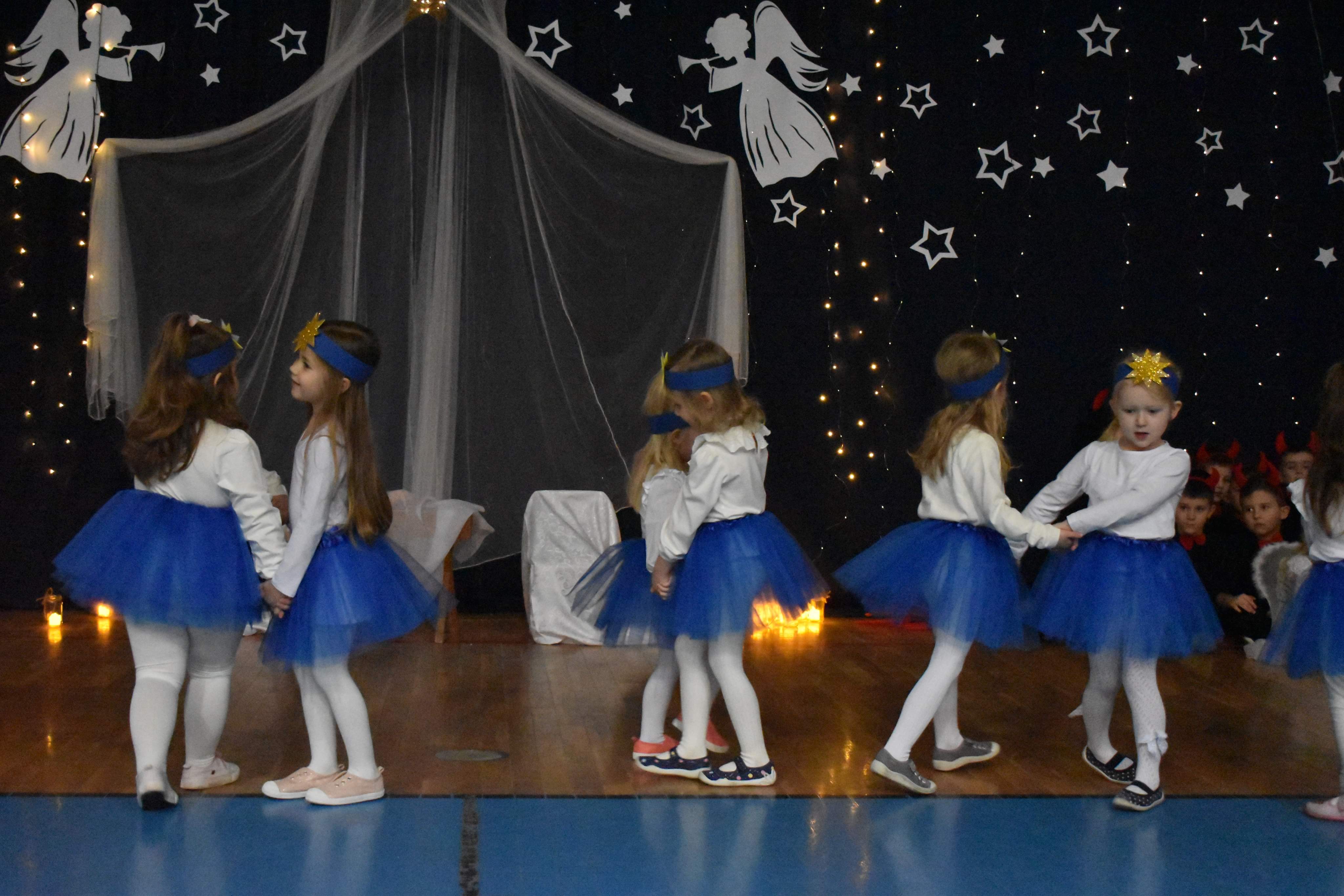 Dziewczynki w białych bluzkach i granatowych spódniczkach i z opaskami gwiazdek na głowach trzymają się za rączki, tańcząc w oarach