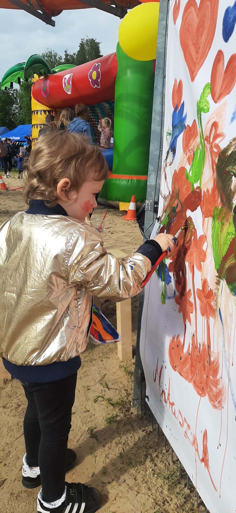 Kilkuletnie dziecko maluje farbami na powieszonych na płocie białych banerach. Za nim widać dmuchane zamki oraz namioty z malowaniem twarzy i pleceniem warkoczy.