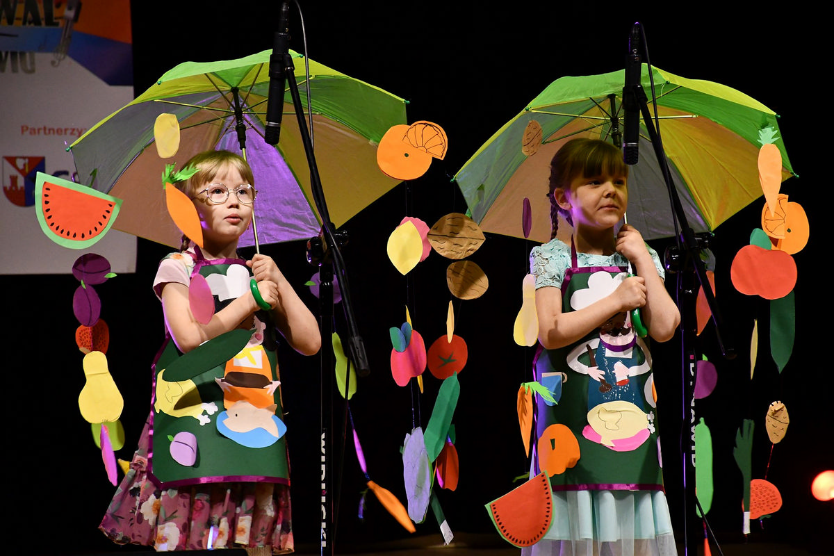 Na scenie dwie dziewczynki w wieku szkolnym z kolorowymi parasolami. Z parasolów zwisają w dół na żyłkach papierowe kolorowe produkty, warzywa i owoce oraz inne promujące zdrową dietę