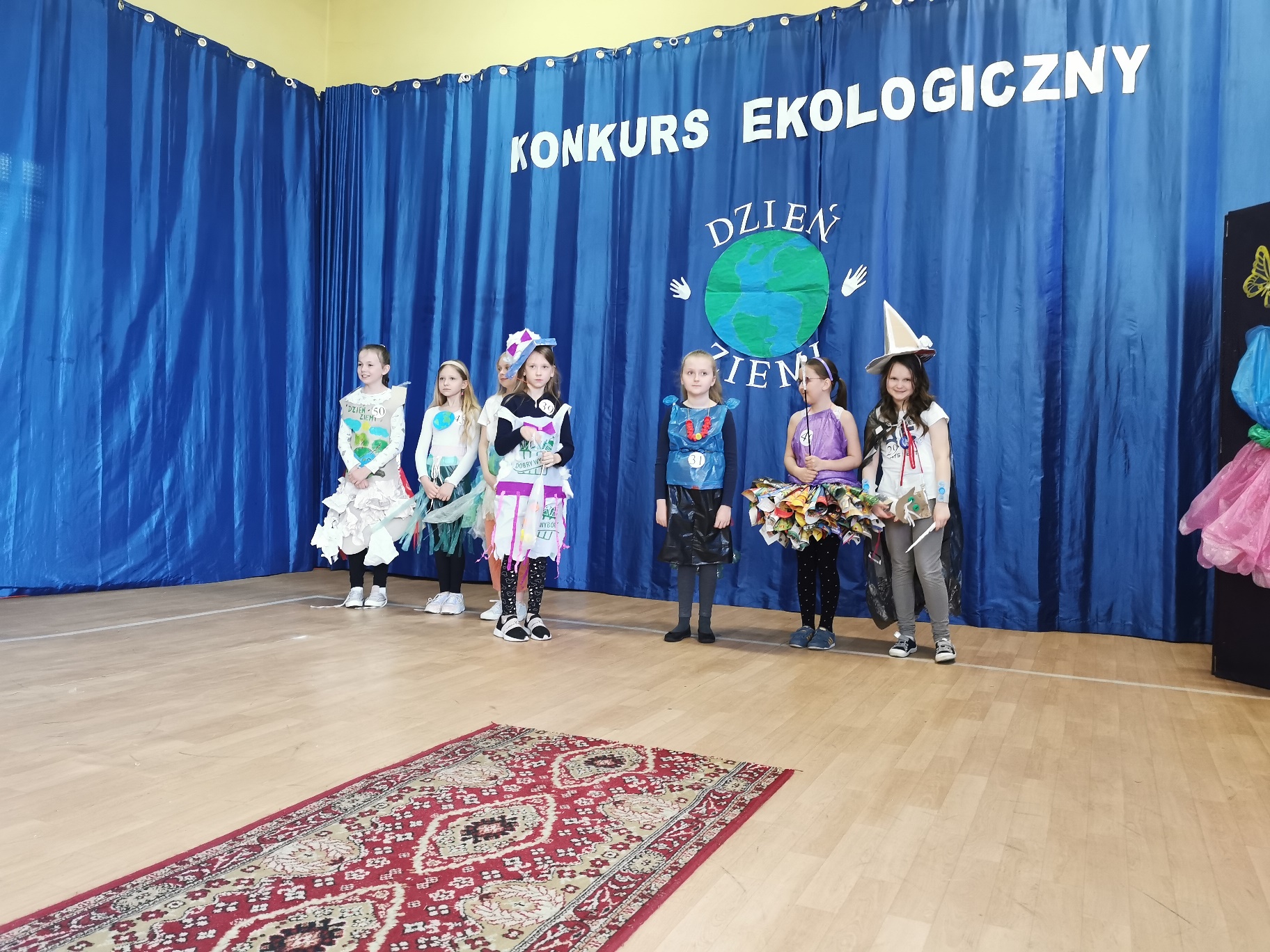 Dzieci w kolorowych strojach stoją przed niebieską kurtyną z napisem Konkurs ekologiczny i obrazem Ziemi