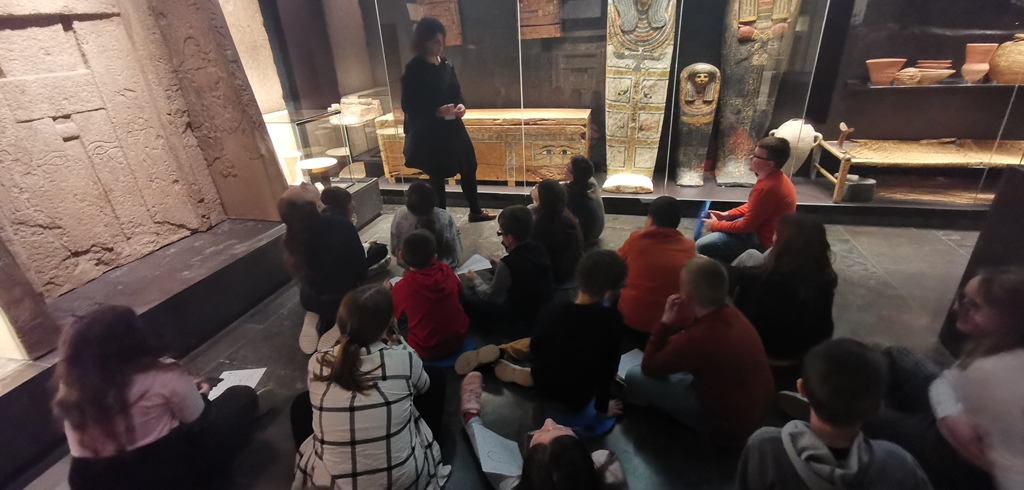 Dzieci siedzą na podłodze sali z przeszklonymi witrynami w których ustawione są sarkofagi. Przemawia do nich kobieta w czarnej sukience