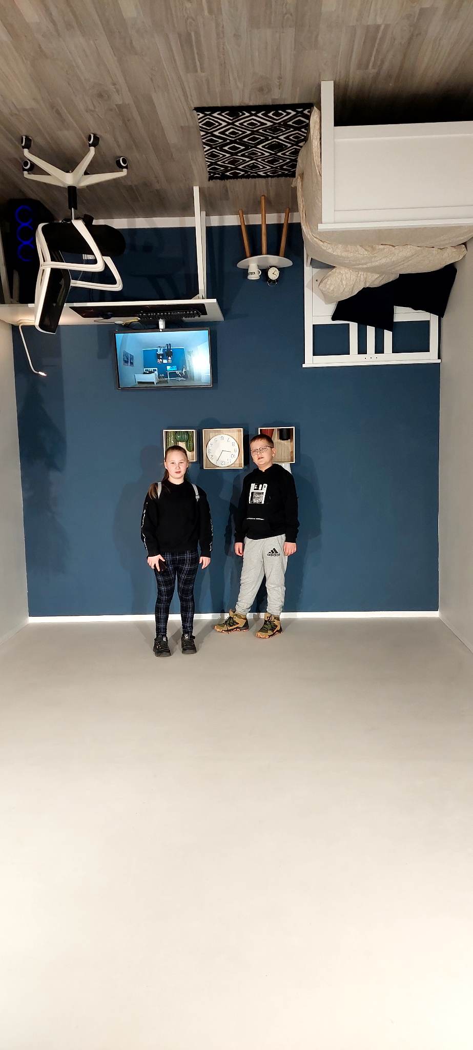 Zdjęcie przedstawia dziewczynkę oraz chłopca, którzy stoją w dziecięcym pokoju obróconym o 180 stopni. 