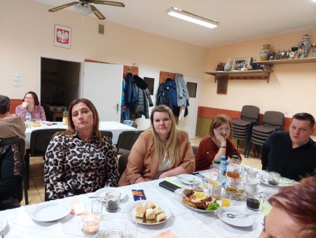 5 kobiet i mężczyzna siedzący przy stole z białym obrusem. Na stole stoją napoje i jedzenie. W tle na ścianie godło Polski, kurtki na wieszakach, puchary na wiszącej na ścianie półce.
