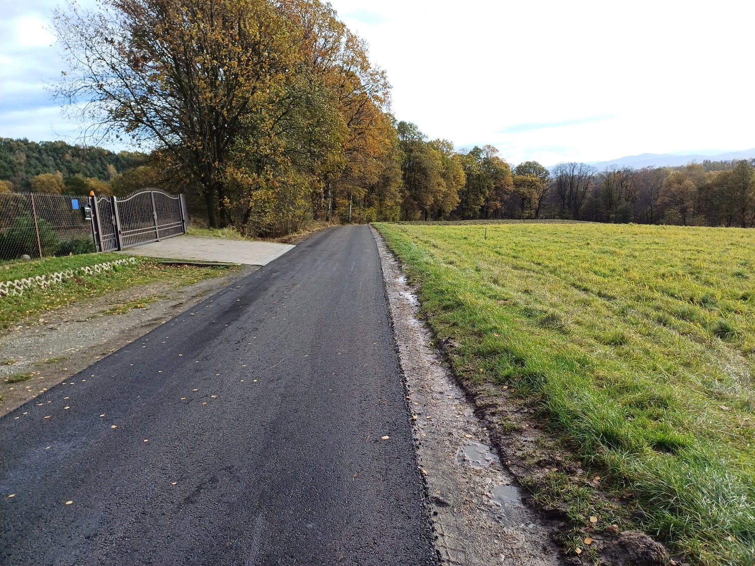 Fotografia przedstawia świeżo wyremontowaną, asfaltową drogę wiejską biegnącą wśród pól i lasu. Po lewej stronie widoczna jest brama wjazdowa na posesję oraz ogrodzenie. Po prawej rozciąga się otwarte pole, a w tle widać drzewa z jesiennym ubarwieniem. Krawędź drogi jest równo wykończona, co wskazuje na zakończone prace remontowe.