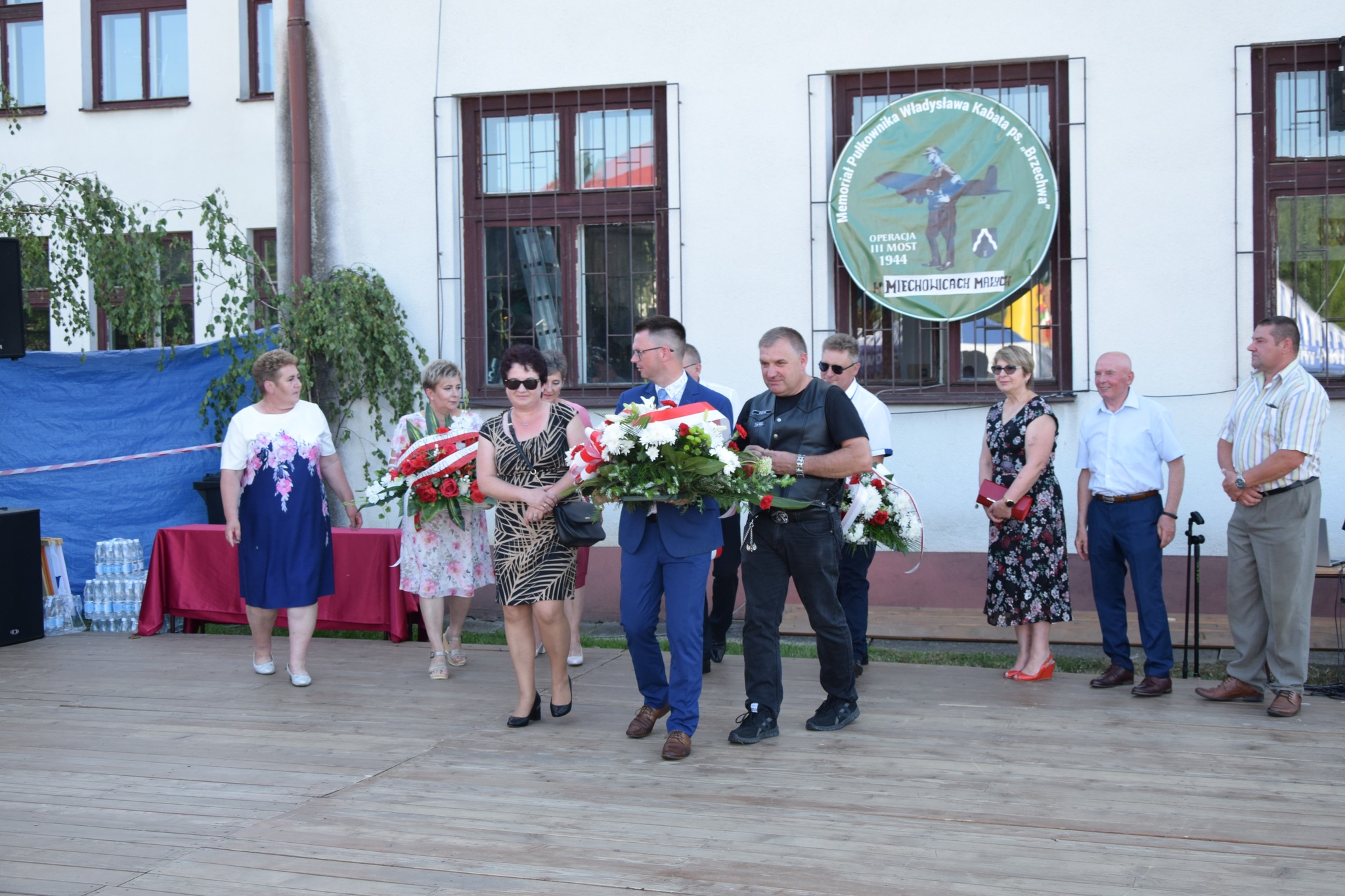 zdjęcie przedstawia delegacje osób, które idą złożyć kwiaty pod pomnikiem i pamiątkową tablicą Płk. Władysława Kabata.