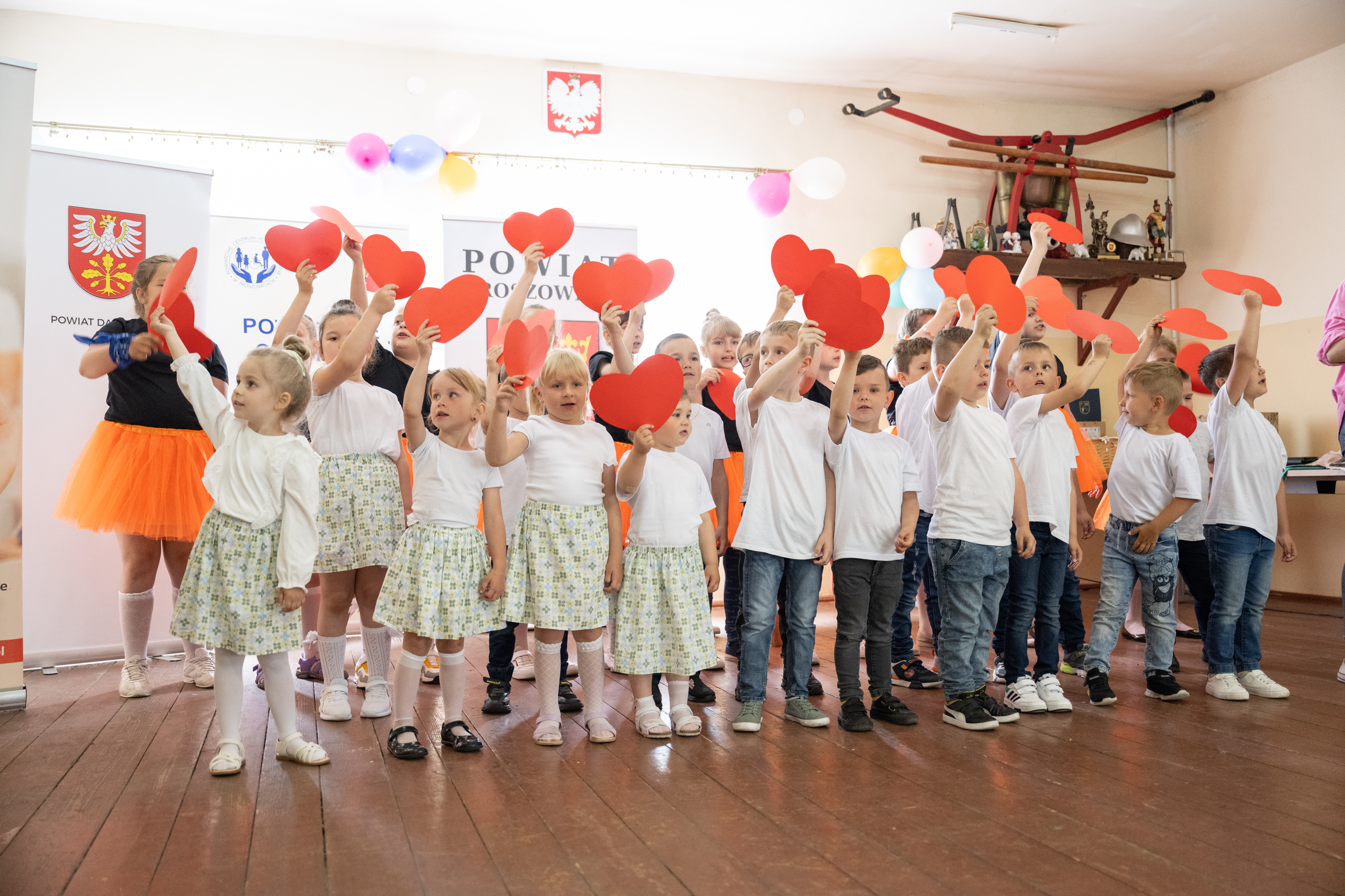 na scenie grupa dzieci chłopców i dziewczynek w wieku przedszkolnym i szkolnym, dzieci trzymają nad głowami papierowe serca w kolorze czerwonym