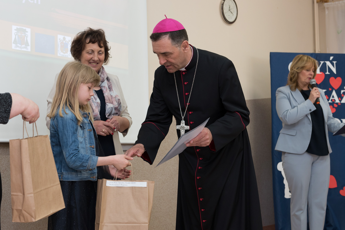 Na fotografii JE ks. Biskup wraz ze starostą sokołowskim wręczają nagrodę jednemu z laureatów. Ks. biskup podaje dziecku torebkę z nagrodami rzeczowymi. 