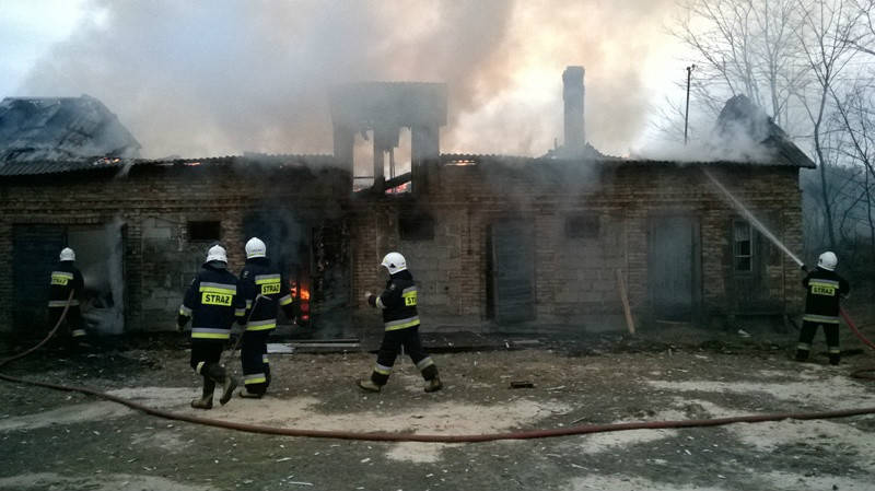 Spalony budynek gospodarczy murowany z białej cegły bez dachu z którego unosi się siwy dym. Przed budynkiem strażacy dogaszający zgliszcza