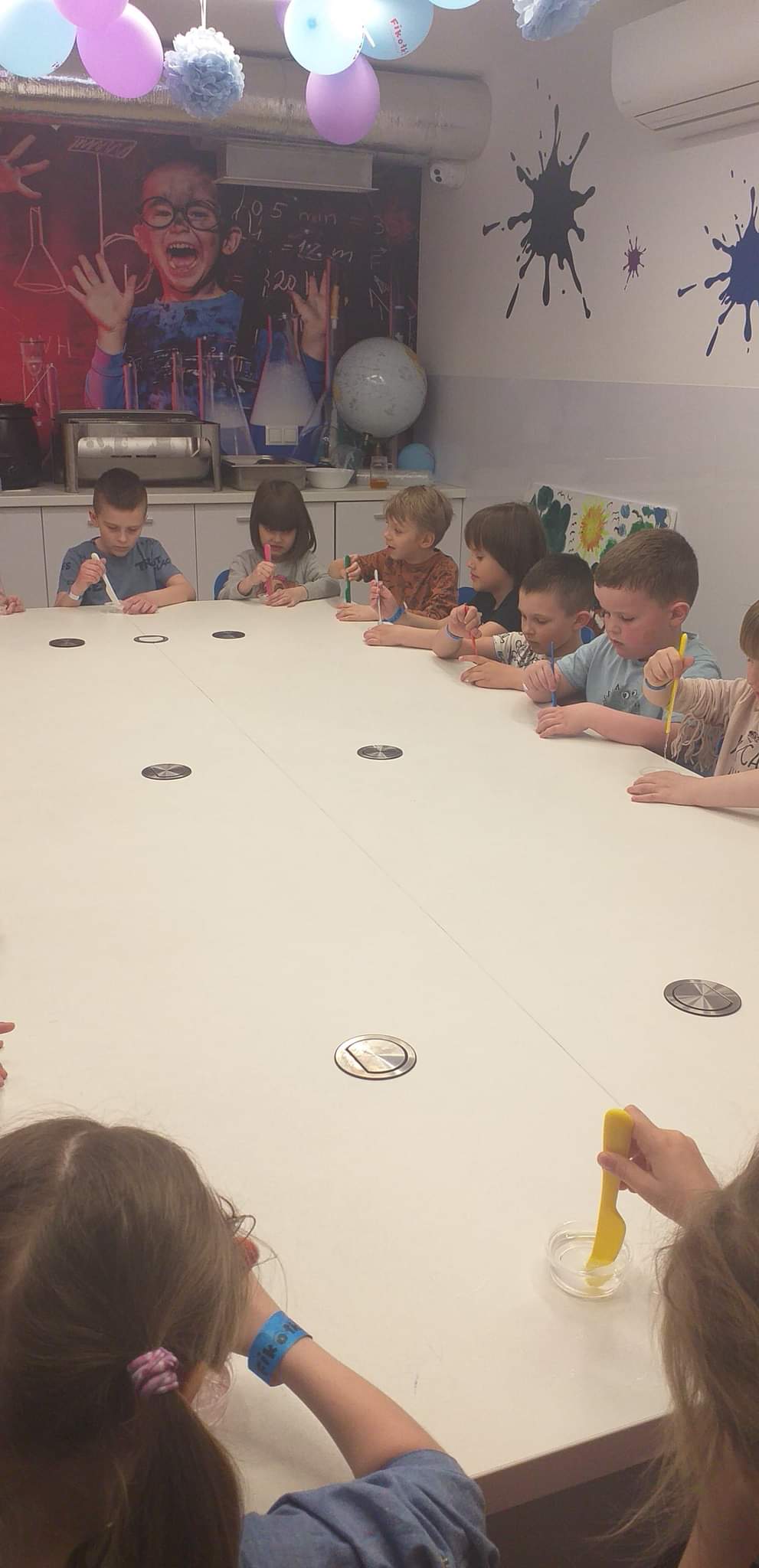 Dzieci siedzą przy długim biały stole. Przed mini leżą plastikowe noże. 