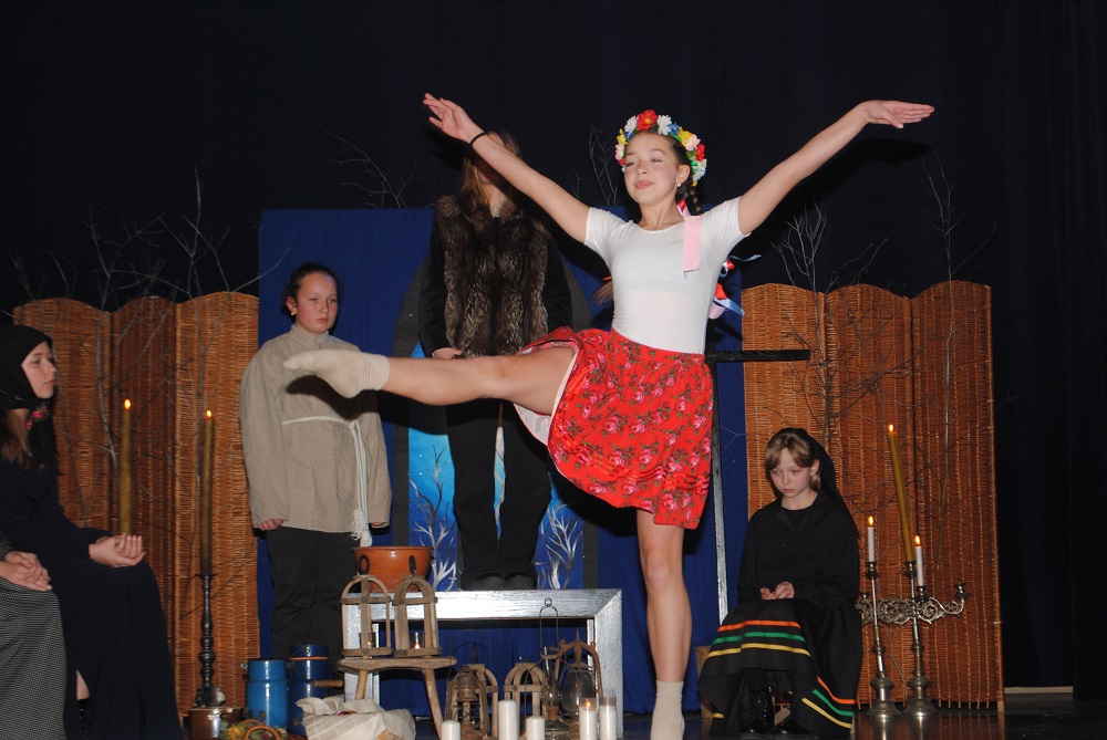 dziewczynka w białej bluzce i czerwonej spódnicy z wiankiem na głowie tańczy na scenie