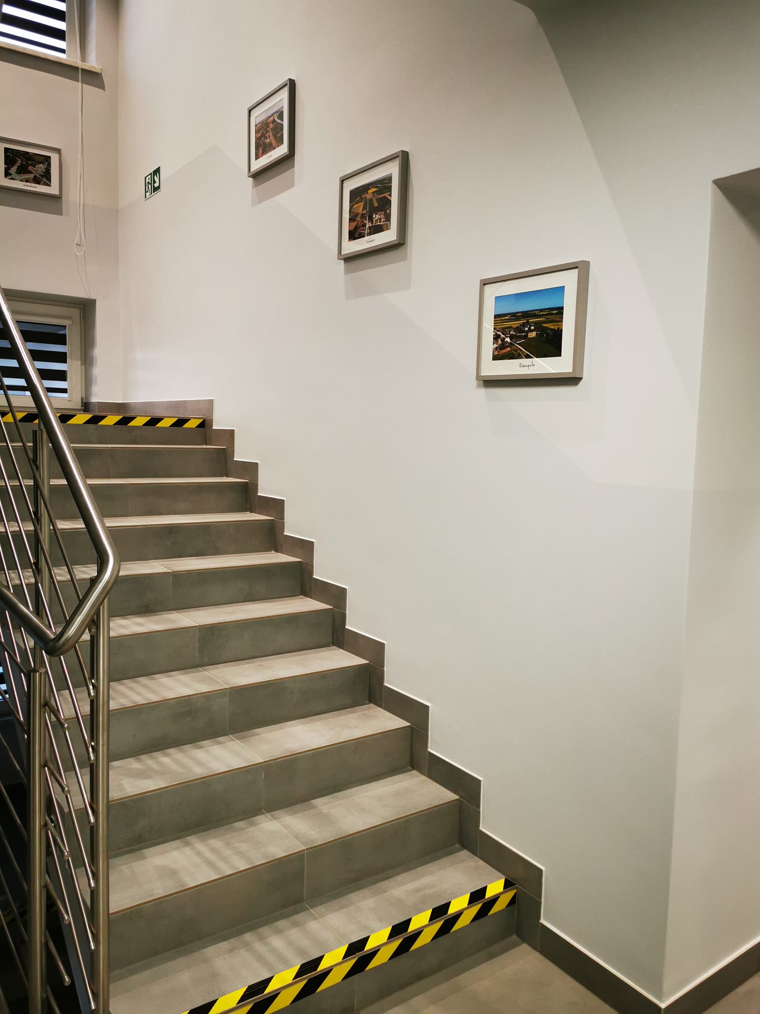zdjęcie przedstawia fragment schodów i ściany w holu budynku urzędu gminy Wietrzychowice. Schody z płytek w kolorze szarym, srebrna bariera. Na ścianach wiszą zdjęcia.