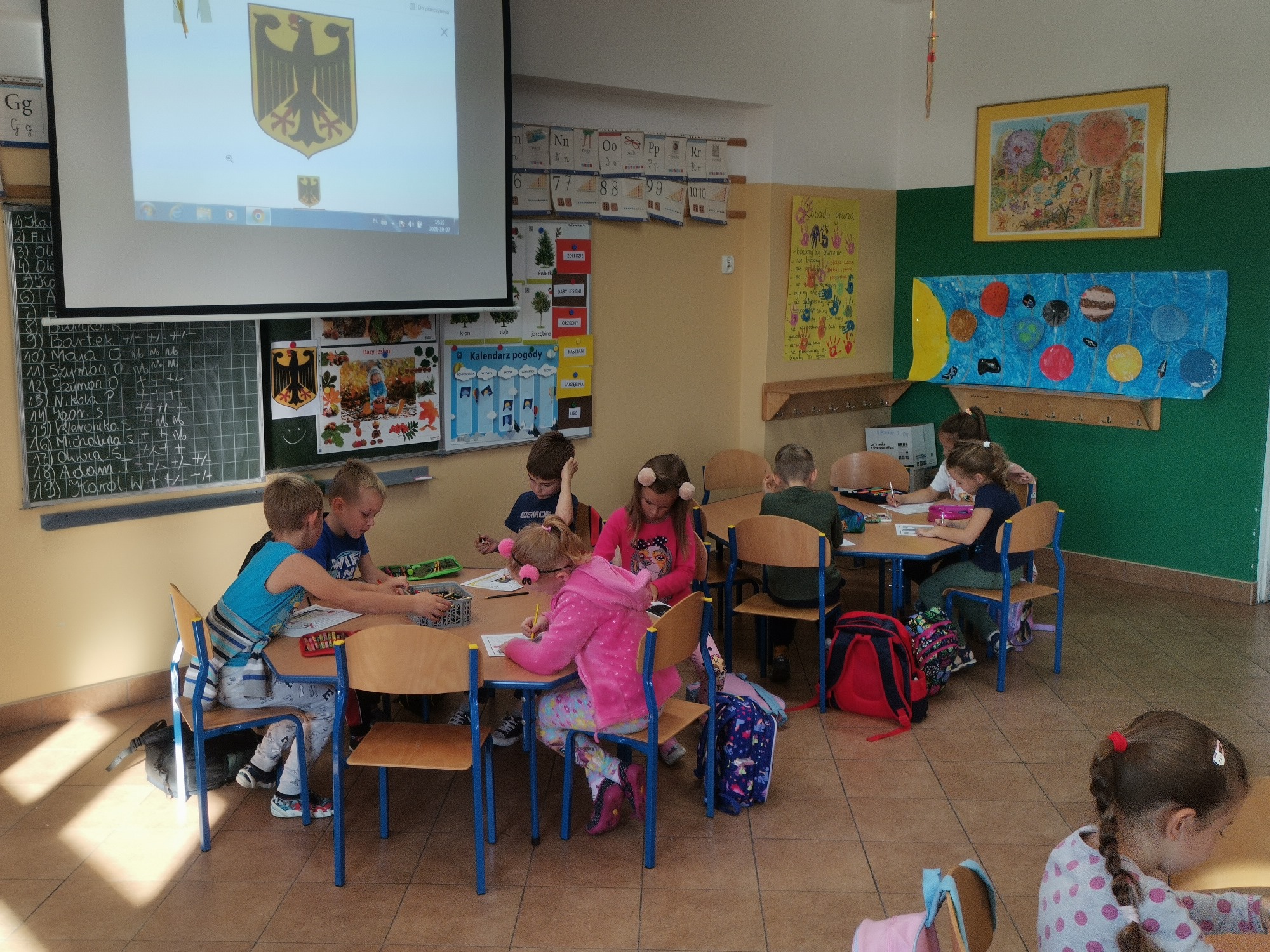 Dzieci siedzą w grupach przy stolikach. Na ekranie przy ścianie wyświetlane jest godło czarnego orła na żółtym tle