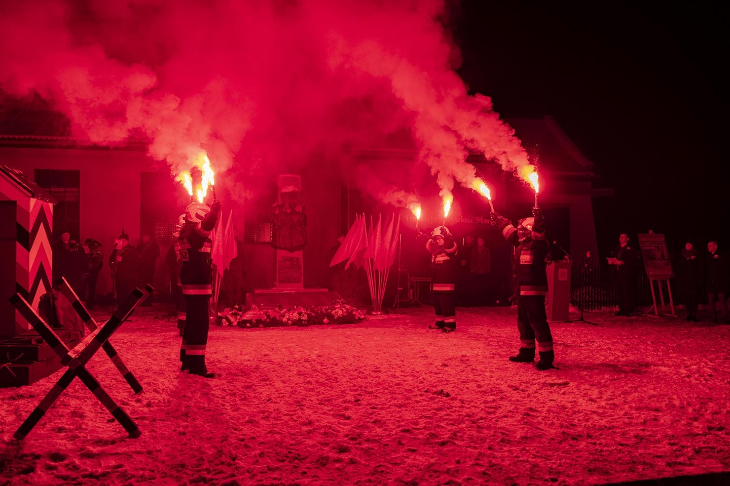 strażacy stojący naprzeciwko siebie, przed pomnikiem Powstańców Wielkopolskich, trzymający race płonące na czerwono