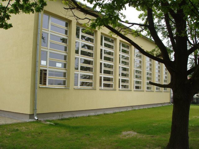 Zółty wysoki budynek z wysokimi dzielonymi oknami. Po prawej stronie rośnie drzewo i rośnie zielona trawa