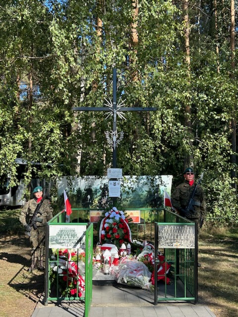Krzyż otoczony zieloną barierką przy nim asysta żołnierzy. Na płycie złożone kwiaty w biało- czerwonych barwach