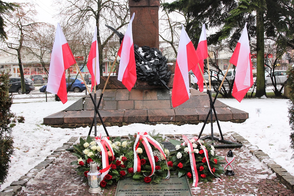 Widok na złożone kwiaty. Na płycie u podstawy monumentu leżą trzy wiązanki z biało - czerwonych róż oraz zapalone 2 znicze. W tle podstawa pomnika z wieńcem laurowym oraz sześć polskich flag