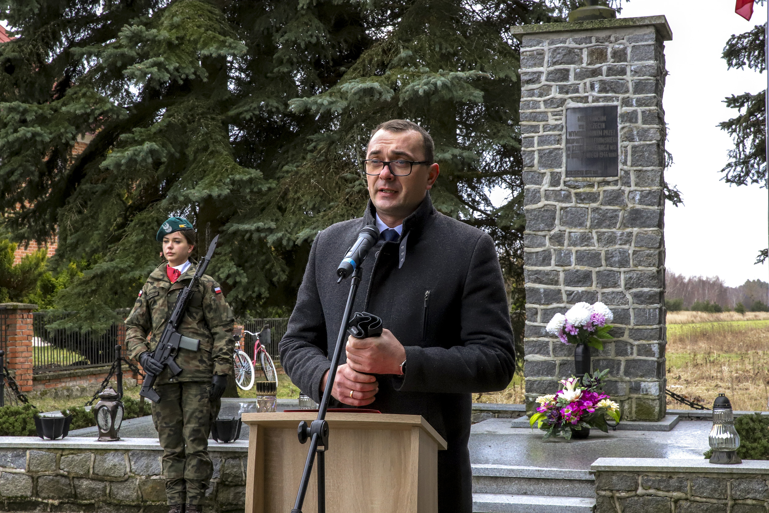 Na zdjęciu Starosta Kraśnicki stojący przy mównicy podczas okolicznościowego przemówienia. W tle za nim znajduje się pomnik upamiętniających pacyfikację z 2 lutego 1944 roku oraz pełniąca staż honorową żołnierka. 