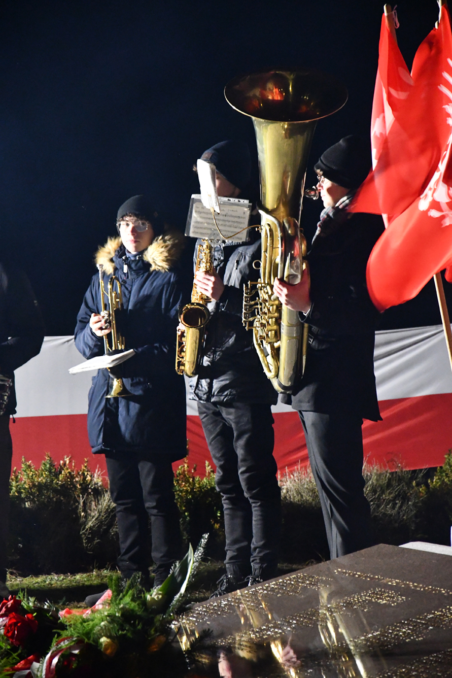 Muzycy grają koncert przy pomniku na saksofonie, trąbce i tubie.