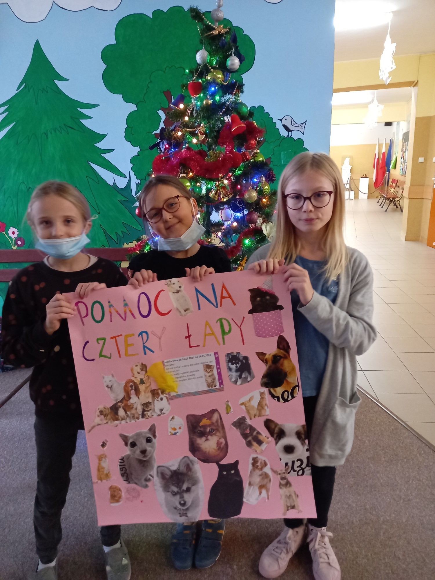Trzy dziewczynki trzymają dużą różową kartkę z napisem Pomoc na 4 łapy z obrazkami psów i kotów