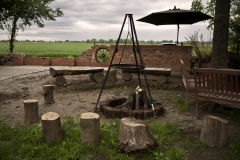 Ułożone w formie ogniska drwa wokoło drewniane stołki do siedzenia w tle parasol ogrodowy oraz zielone pola