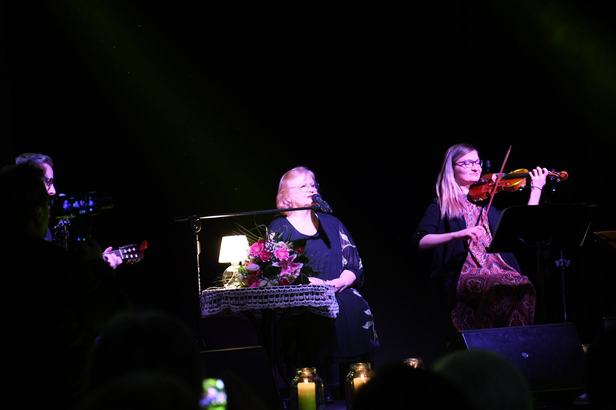 Trzy osoby siedzące na scenie, Stanisława Celińska oraz dwoje muzyków, kobieta grająca na skrzypcach i mężczyzna grający na gitarze. Przy Stanisławie stolik z lampką. Zdjęcie zrobione w trakcie koncertu.