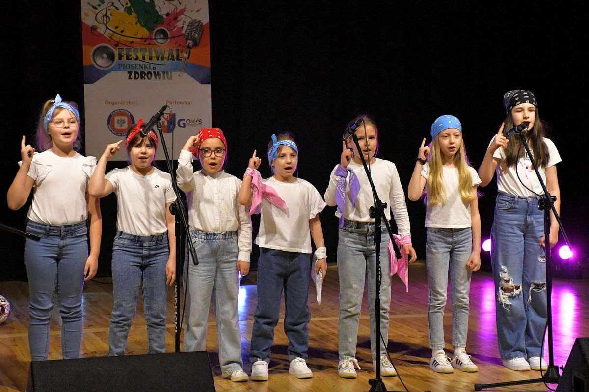 Na zdjęciu na oświetlonej scenie z drewnianą podłogą widzimy jedno z wykonań konkursowych. Piosenkę wykonuje siedem dziewczynek w wieku szkolnym na głowach założone mają kolorowe chustki.