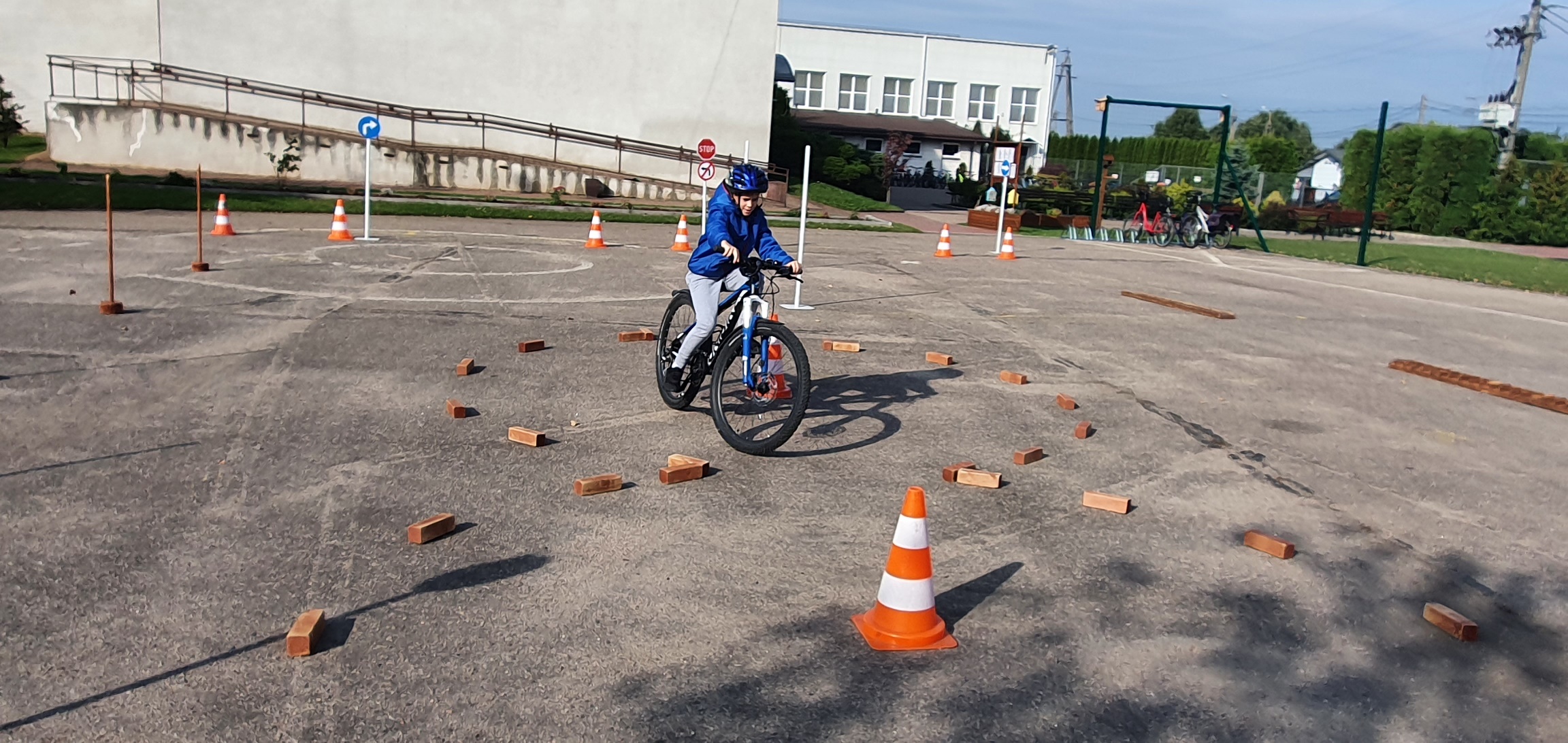 Chłopiec w niebieskiej kurtce i niebieskim kasku jedzie rowerem po placu, gdzie trasę jazdy wyznaczają cegły i pachołki