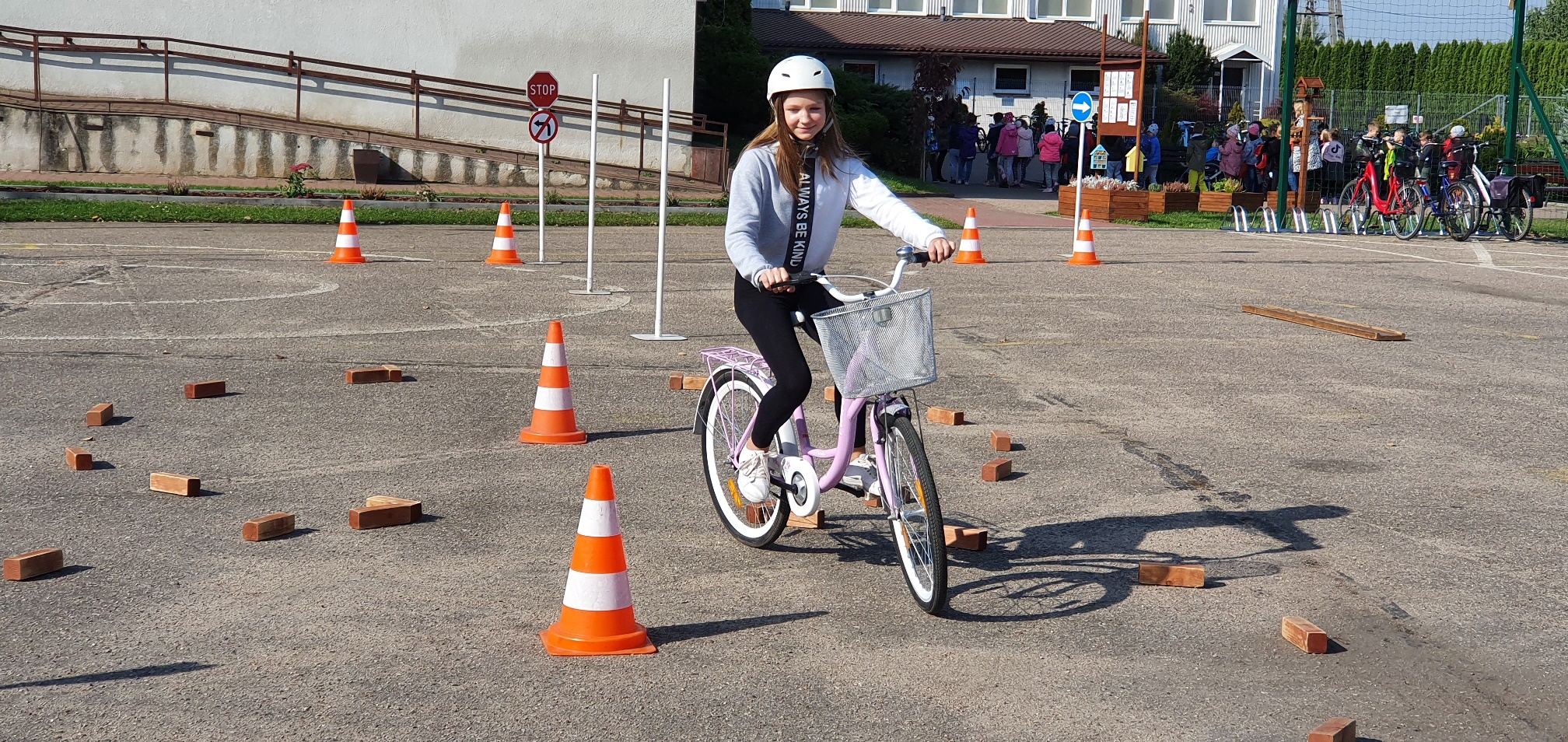 Dziewczynka w białym kasku jedzie rowerem po placu, gdzie trasę jazdy wyznaczają cegły i pachołki