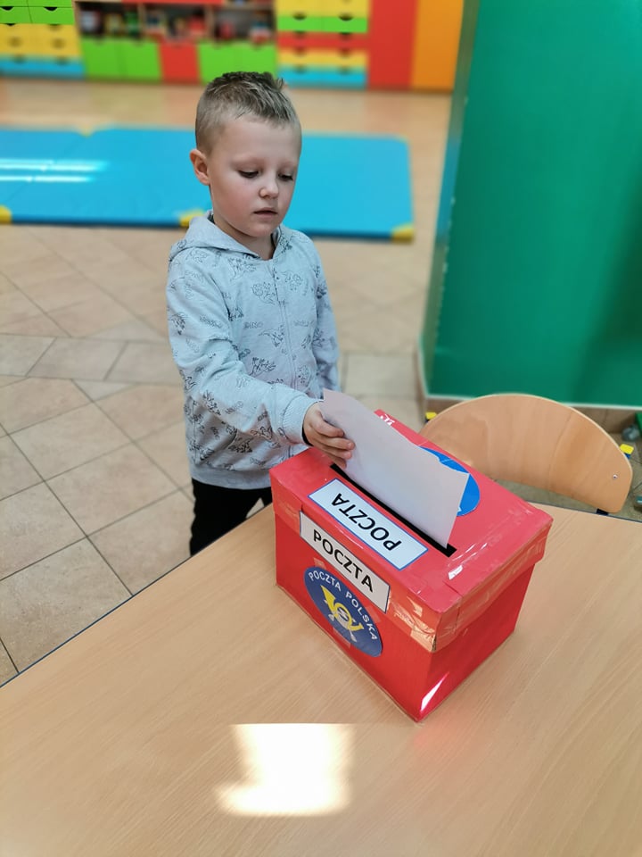 Chłopiec wkłada kartkę do czerwonej skrzynki z napisem Poczta