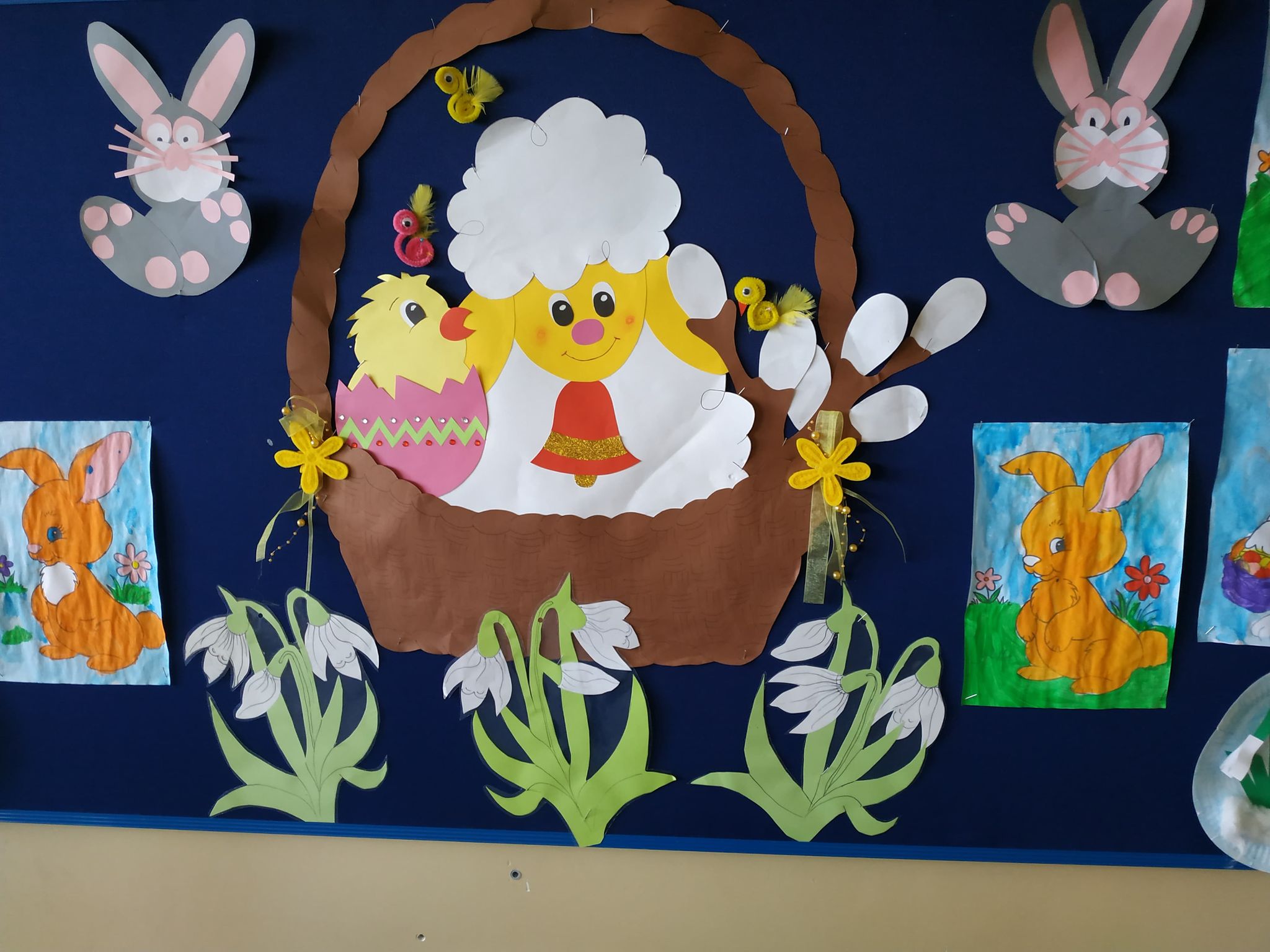 Gazetka szkolna z obrazkiem koszyczka z barankiem, kurczaczkiem i baziami. Po bokach obrazki króliczków a pod nimi kwiaty