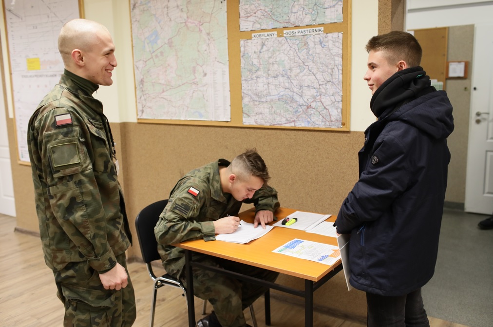 zdjęcie przedstawia stojącego i uśmiechającego się żołnierza oraz młodego chłopaka w pomieszczeniu, obok przy stoliku siedzi i pisze drugi żołnierz. Na ścianie wisi tablica na której zawieszone są mapy.