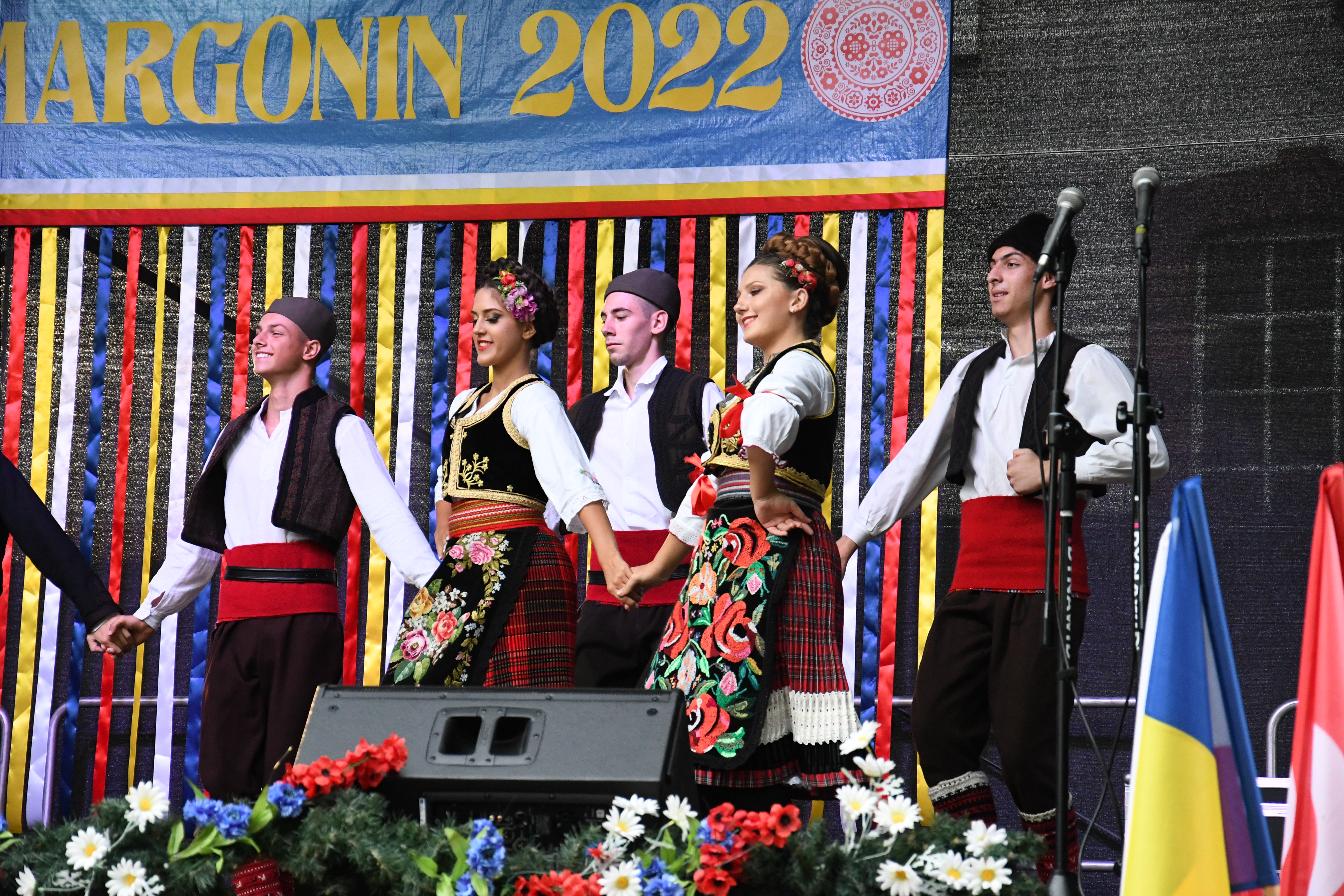 Zespół Zavicaj z Serbii na scenie w komplecie ok. 5 osób, dziewczęta z przodu i chłopcy z tyłu. Ubrani w tradycyjne stroje ludowe. Z tyłu sceny wielki baner z napisem InterFolk Margonin 2022 oraz na pierwszym planie flagi krajów biorących udział w festiwalu.