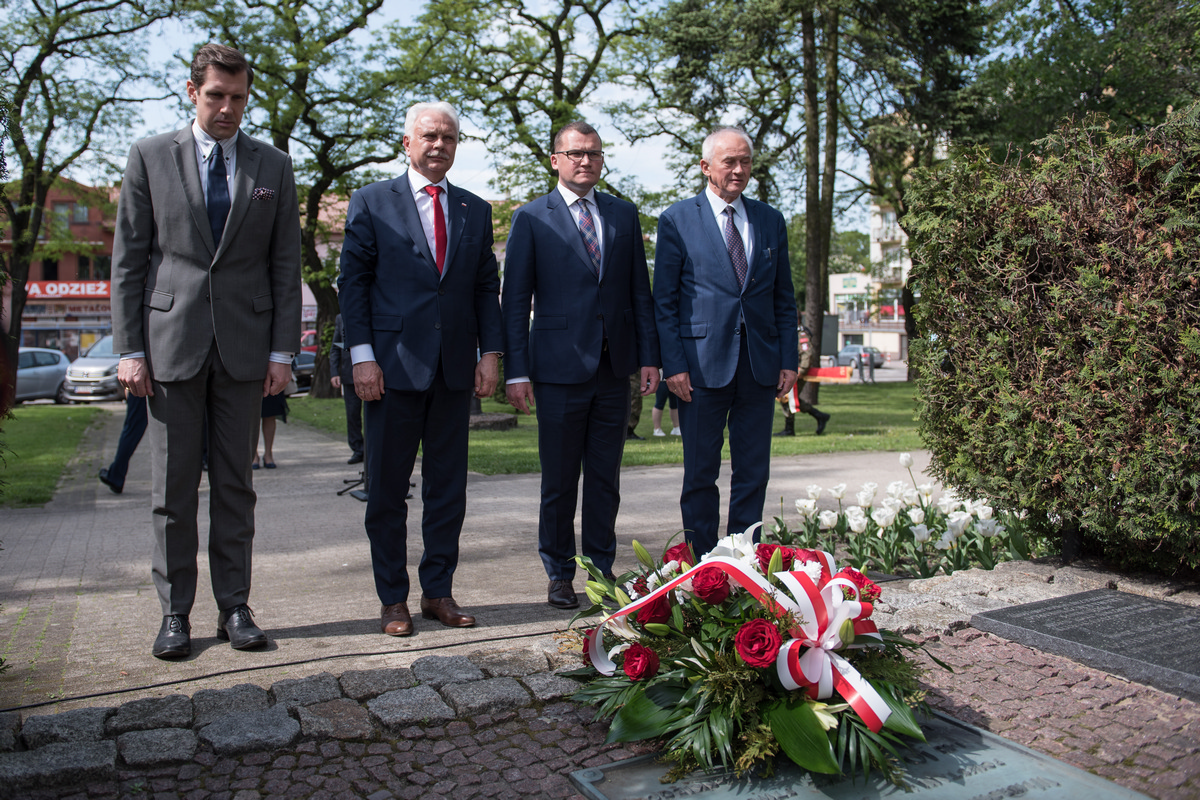 Moment po złożeniu kwiatów pod pomnikiem ks. Brzóski w Sokołowie Podlaskim. W pierwszym planie leżąca u podstawy pomnika wiązanka z róż z biało - czerwoną kokardą. W drugim planie delegacja rządowa. W tle park.