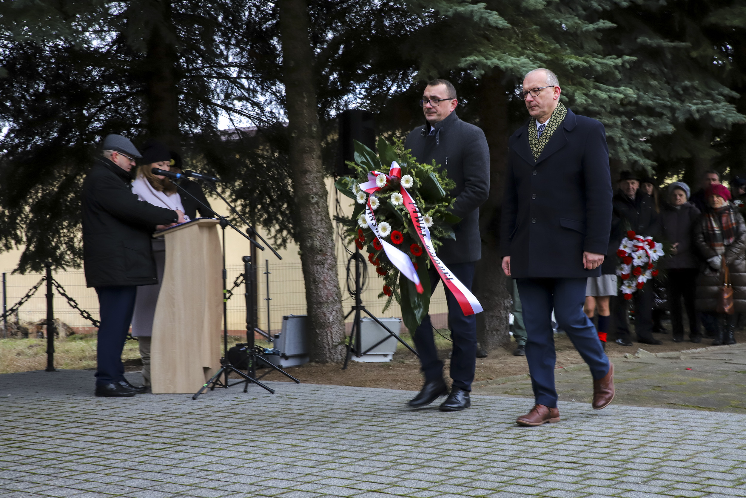 Zdjęcie przedstawia moment składania wieńców przy pomniku. Starosta Kraśnicki oraz Członek Zarządu zmierzają z wieńcem w kierunku pomnika. W tla za nimi znajdują się inni uczestnicy obchodów.
