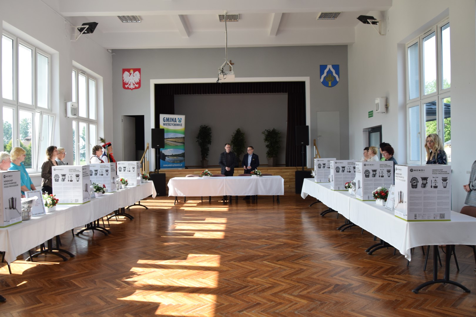 zdjęcie przedstawia dwa stoły ułożone wzdłuż sali widowiskowej Gminnego Centrum Kultury przy których zgromadzone są KGW, na stołach stoją sprzęty