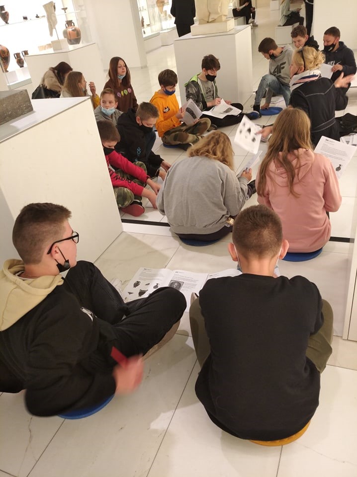 Dzieci siedzące na białej podłodze trzymają kartki z tekstem. Obok nich stoją małe kolumny ze szklanymi kloszami