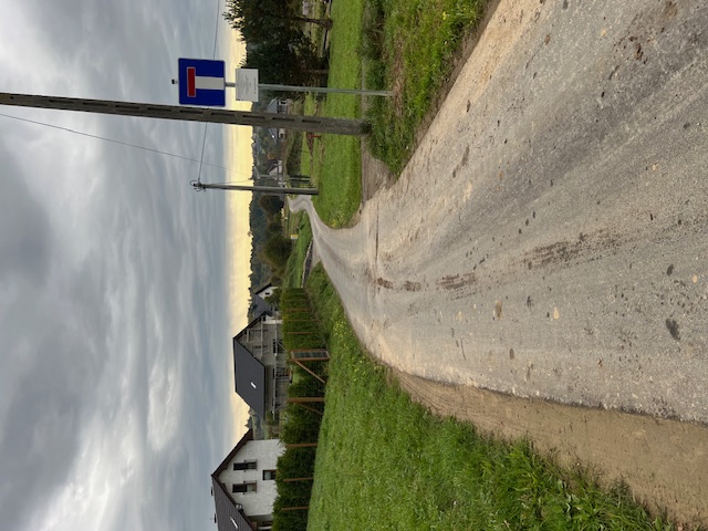 Zdjęcie przedstawia wyremontowaną asfaltową, wąską drogę wiejską, biegnącą przez teren o zabudowie jednorodzinnej i polach uprawnych. Droga jest świeżo utwardzona i estetycznie wykończona. Widać domy w budowie i tereny zielone. 