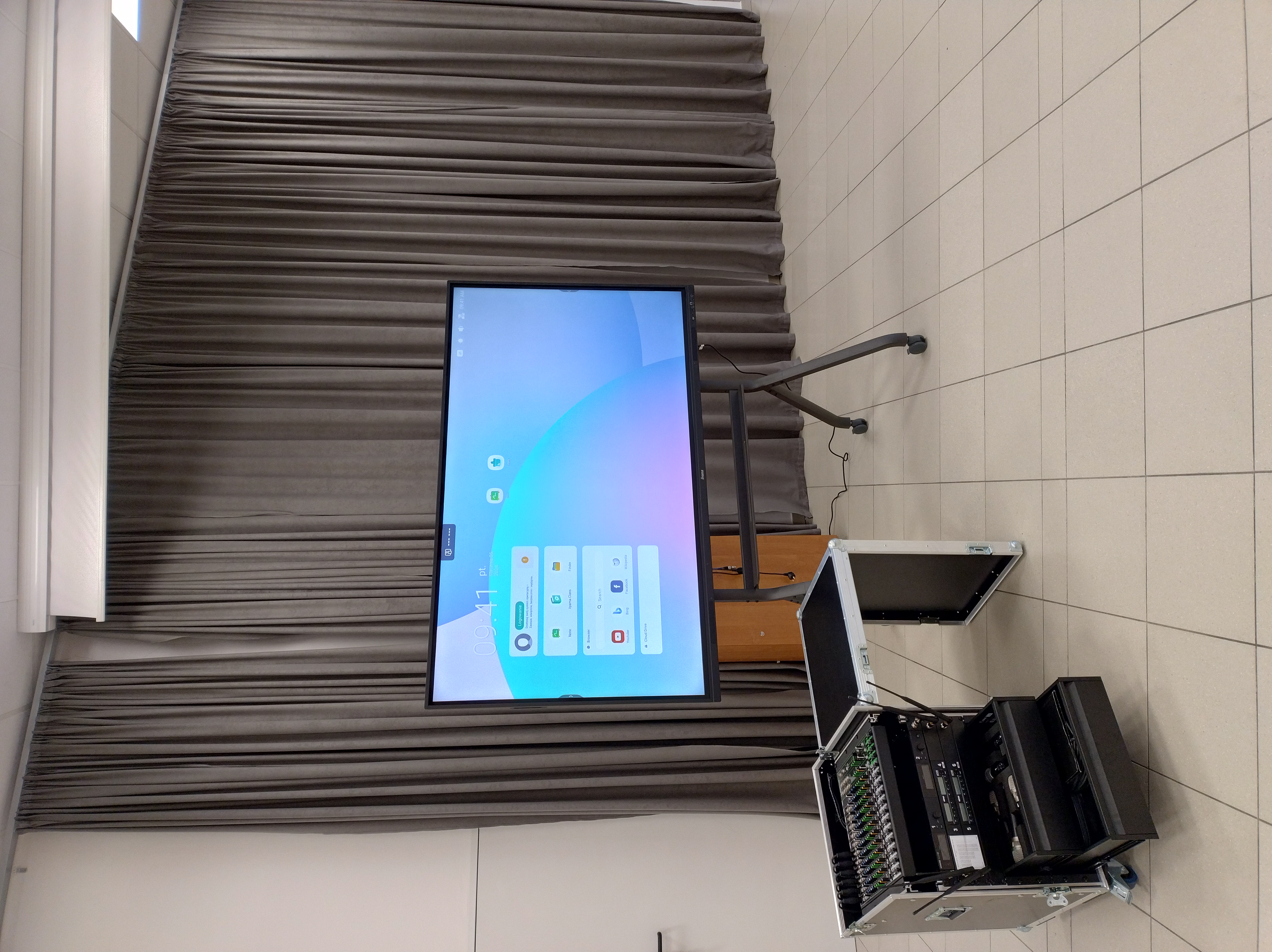 Zdjęcie przedstawia nowoczesny monitor interaktywny na mobilnym statywie z podłączonym zestawem multimedialnym. Obok znajduje się otwarta walizka typu rack z mikserem audio, odbiornikami i mikrofonami bezprzewodowymi.