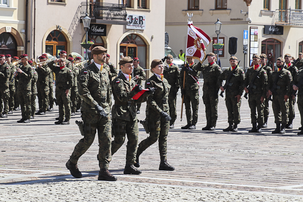 zdjęcie przedstawia w tle salutujących żołnierzy, dwoje mężczyzn i jedna kobieta idą, żołnierz w środku trzyma w rękach złożoną flagę Polski.