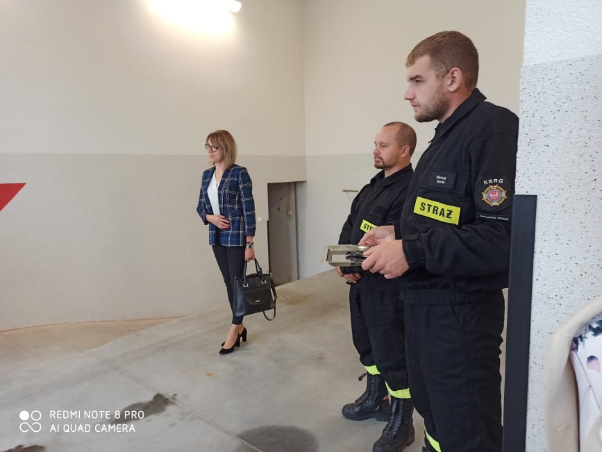 Od prawej dwóch strażaków ubranych w mundury bojowe, w dalszej odległości Pani Monika Bochenek Król. Osoby ze zajęcia stoją w środku budynku OSP Jadowniki Mokre