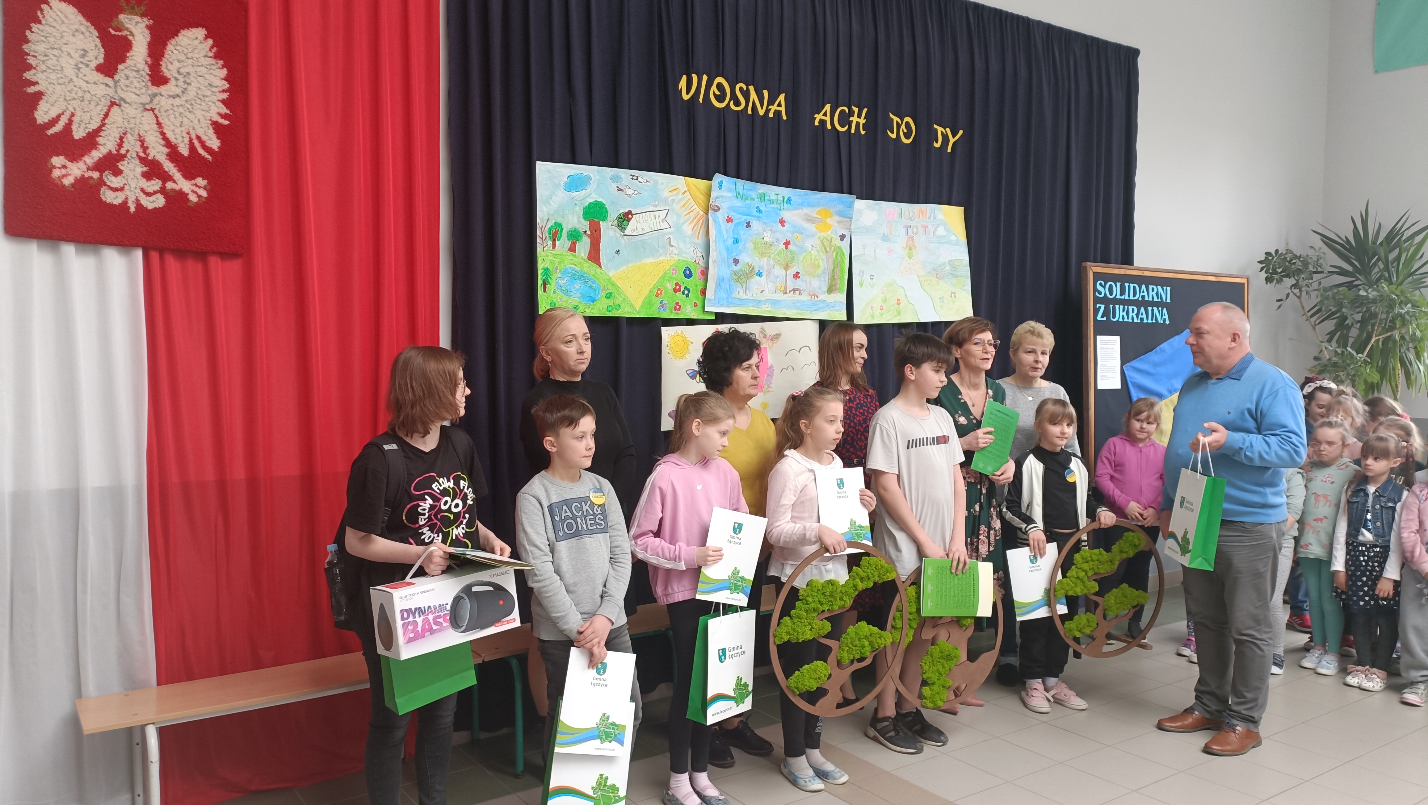 Zdjęcie przedstawiające dzieci i dorosłych na wręczeniu nagród w konkursie, w tle Godło Polski, tekst: " Wiosna Ach To Ty" pod nim narysowane pejzaże wiosny