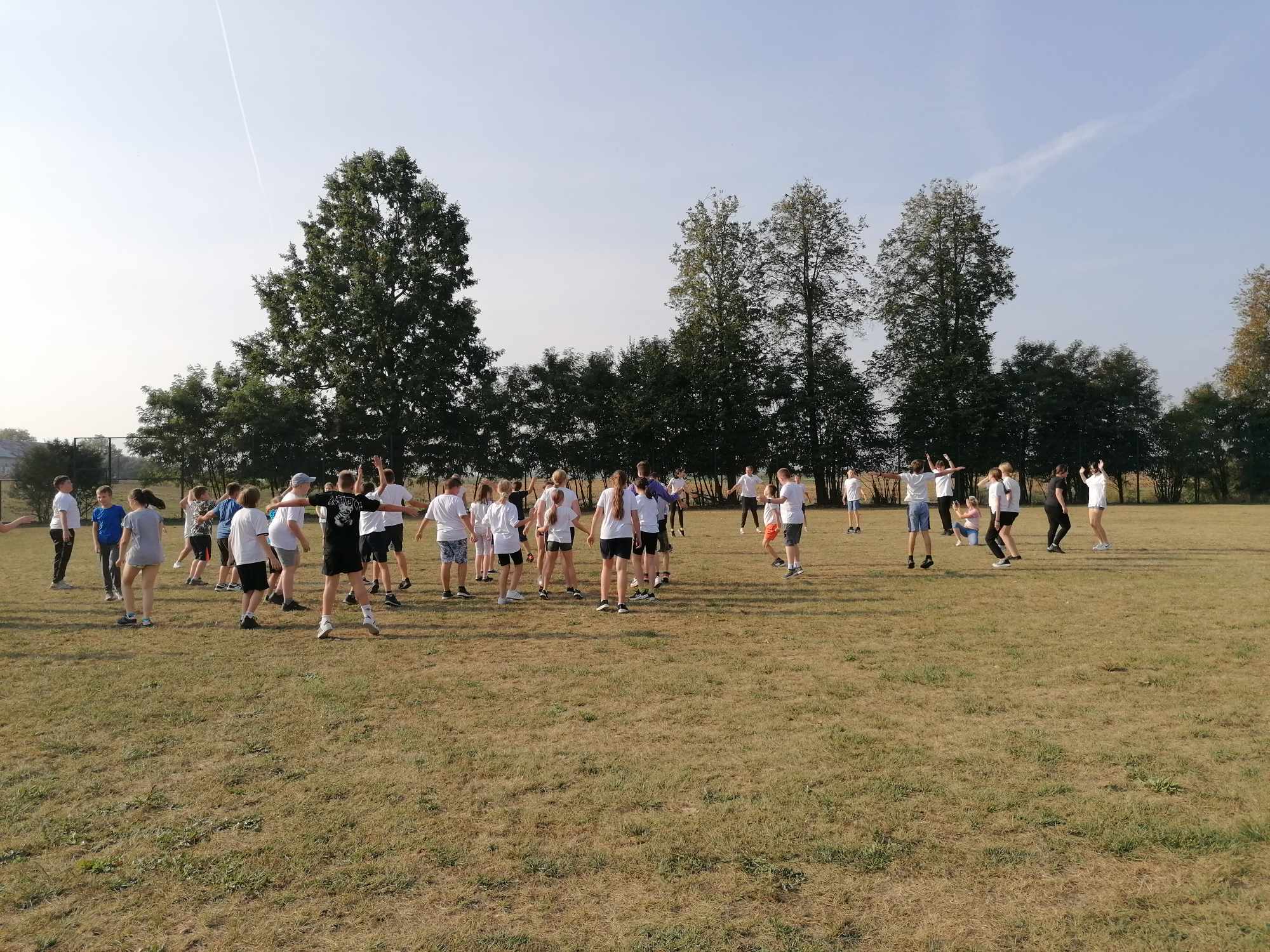 Grupa uczniów ubranych na sportowo wykonuje rozgrzewkę na trawiastym boisku szkolnym.
