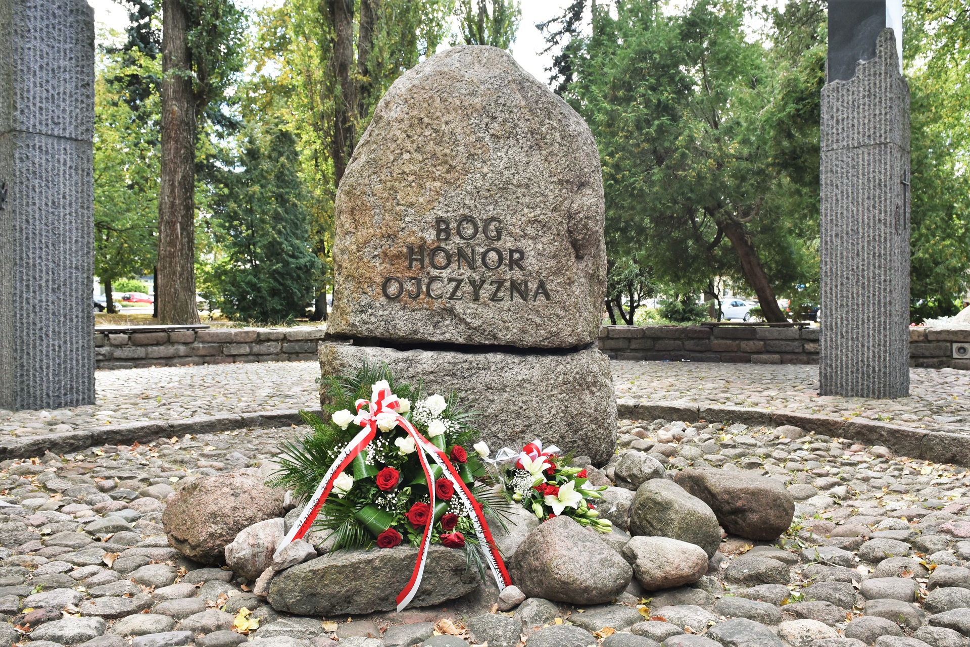 Na zdjęciu kamień w centralnej części pomnika Czynu Niepodległościowego w Sokołowie Podlaskim. Na kamieniu napis Honor, Bóg, Ojczyzna. U podnóża kamienia widać ułożone dwie wiązanki z biało - czerwonych kwiatów.