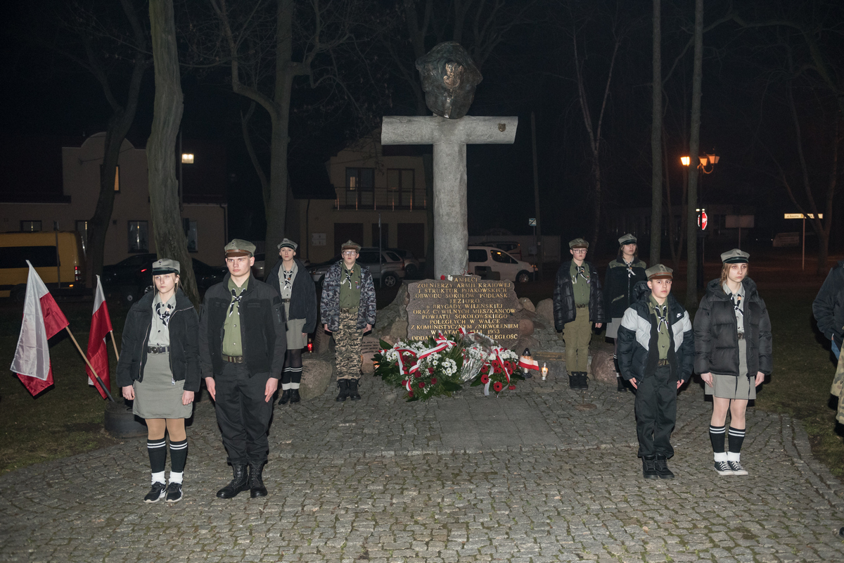W wieczornej scenerii skweru NMP w Sokołowie Podlaskim widzimy frontalną fotografię pomnika z ustawioną po jego lewej i prawej stronie wartą honorową złożoną z sokołowskich harcerzy w mundurach.