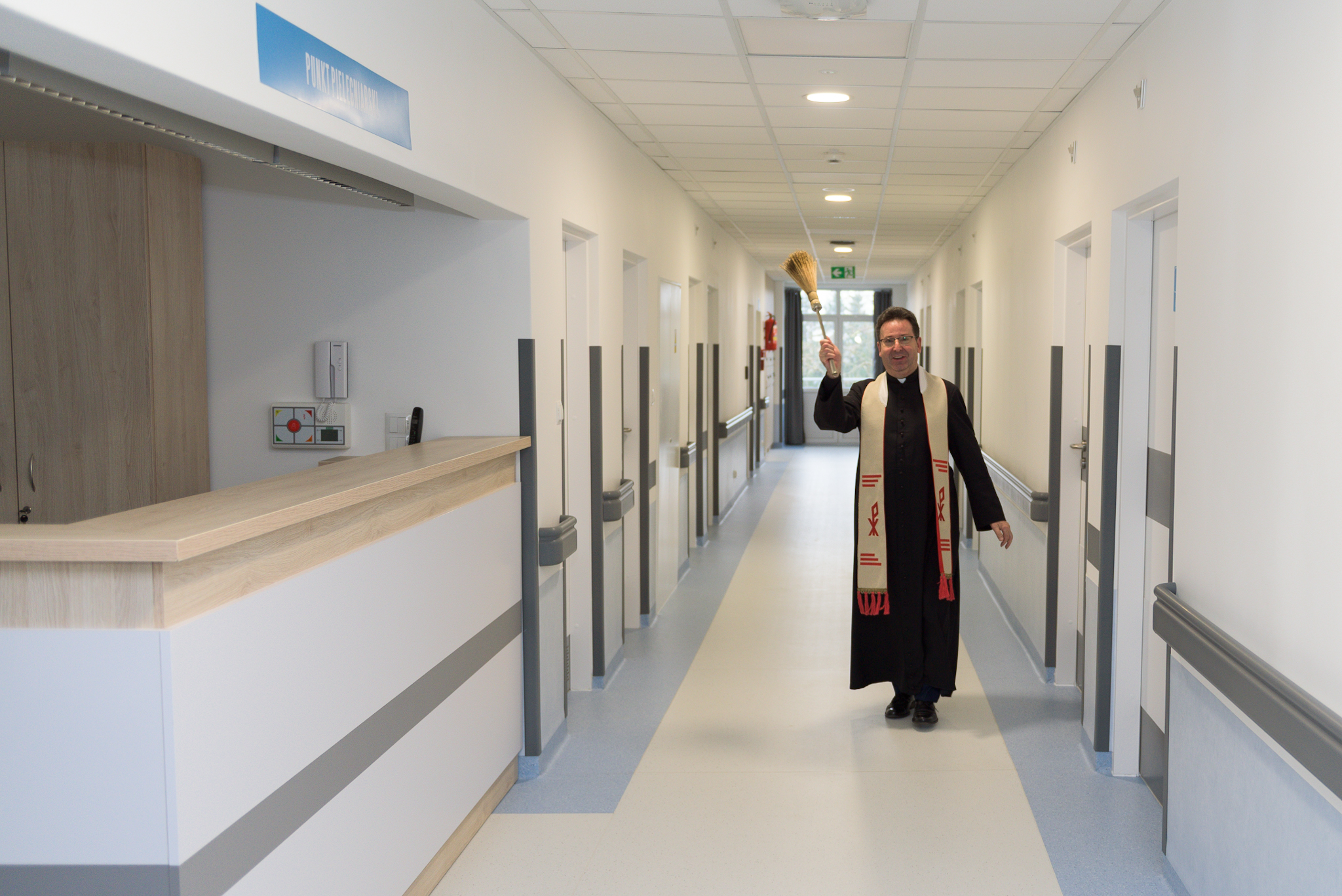 Widok na punkt pielęgniarski oraz korytarz przez który idzie ksiądz i święci wodą święconą kolejne pomieszczenia.