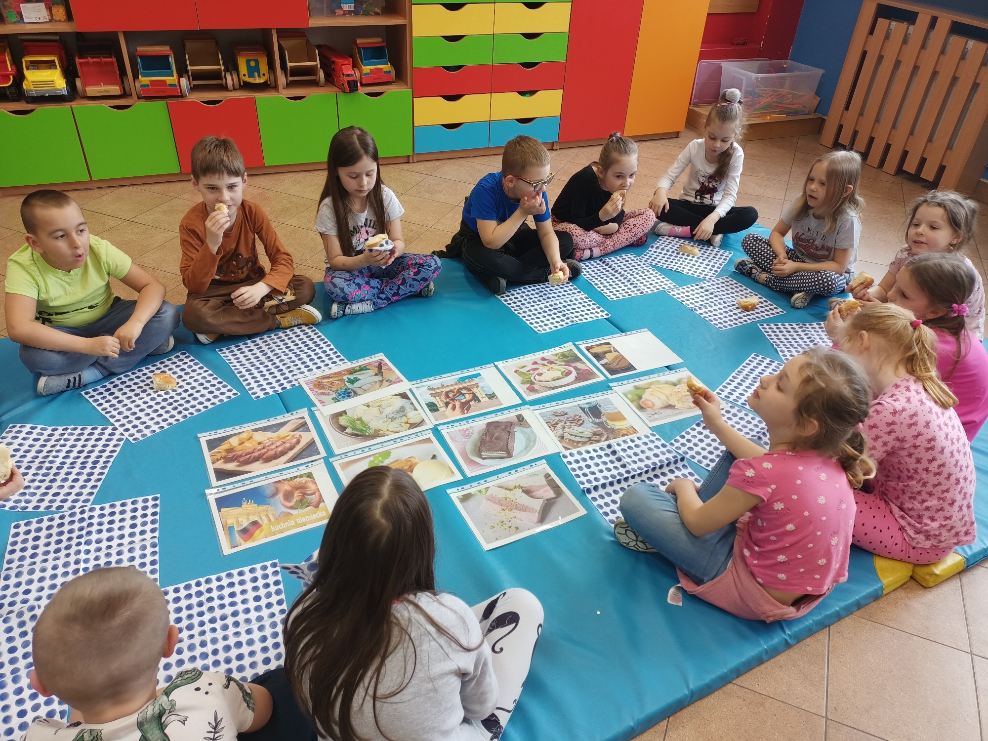 Dzieci siedzące w okręgu na niebieskim materacu. Przed nimi leżą kolorowe obrazki z potrawami