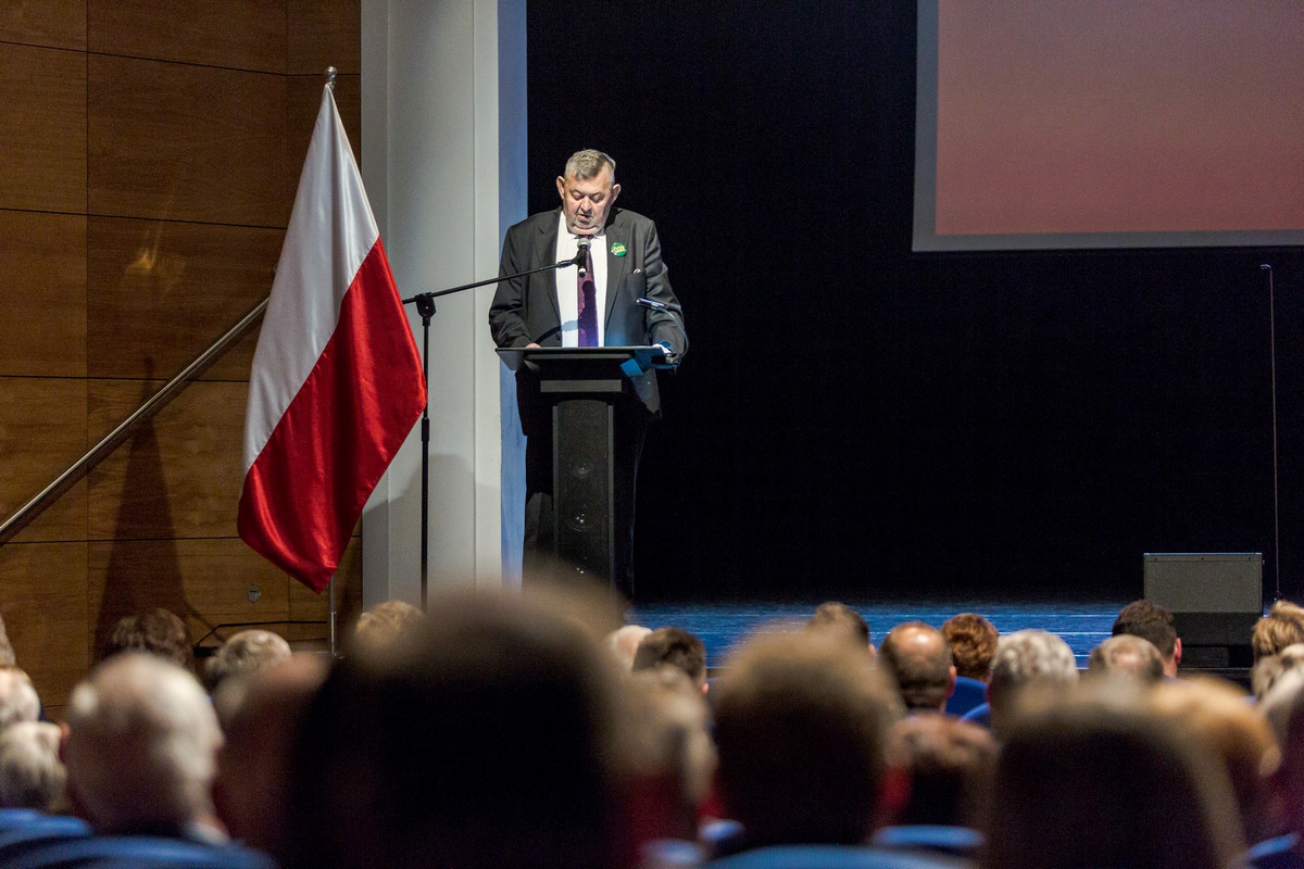 Widok z perspektywy widowni na scenę. Po lewej stronie ustawiona biało - czerwona flaga na statywie w kolorze srebra. Na środku kadru za ciemną mównicą przemawia burmistrz miasta Sokołów Podlaski Bogusław Karakula, który wygłasza prelekcję na temat historii samorządu.